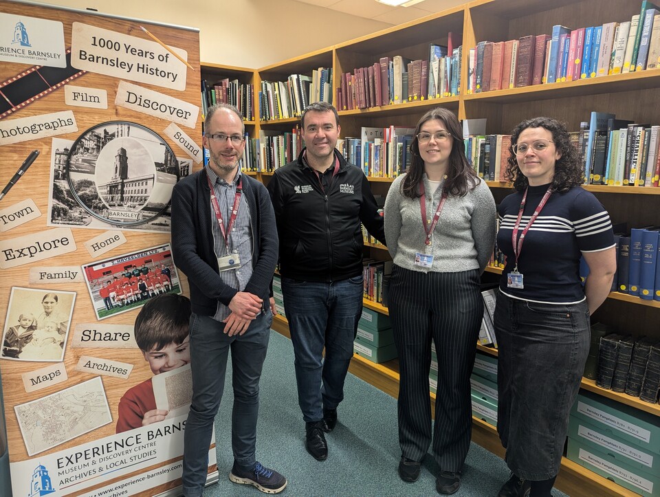 Four people stand in a library next to a banner that reads "Experience Barnsley: Archives & Local Studies." Shelves filled with books are visible in the background.